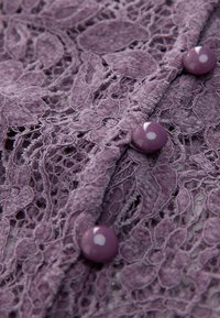 Gros plan sur un tissu en dentelle violette avec des motifs floraux ornés et trois boutons violets brillants le long d'une couture.