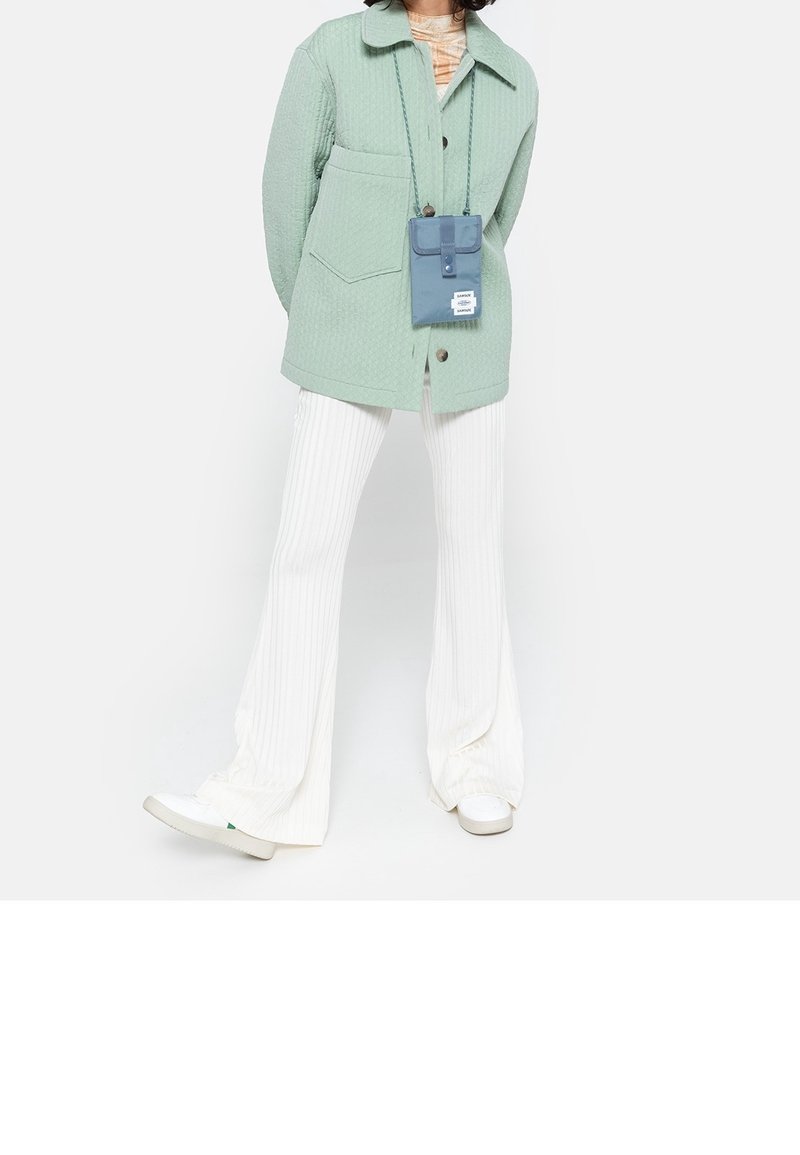 Giacca imbottita verde chiaro con collo, abbinata a pantaloni bianchi a coste svasati e scarpe da ginnastica bianche. È presente una piccola borsa a tracolla blu.