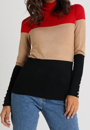 Femme portant un pull en couleurs bloc rouge, beige et noir, associé à un jean bleu, posant avec une main près de sa hanche.