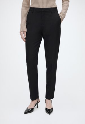 Femme portant un pantalon noir slim, un pull côtelé beige et des talons slingback noirs à bout pointu, debout devant un fond blanc.