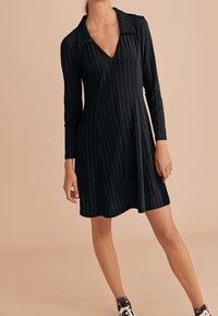 Robe noire à manches longues avec un décolleté en V, dotée de rayures verticales et d'une jupe légèrement évasée, portée avec des baskets noir et blanc.