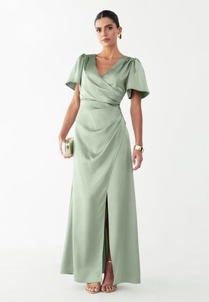 Donna che indossa un abito avvolgente in raso verde chiaro con maniche svolazzanti e spacco frontale, che tiene una piccola borsa clutch dorata con gioielli dorati e sandali.