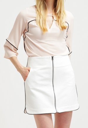 Žena ve světle růžové blůze s černým lemováním a bílé sukni s předním zipem a černým okrajem, stojí s rukou v kapse.