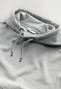 Sudadera gris con cordón, textura suave, cuello doblado y una etiqueta que dice "NEXT". Tejido liso con costuras mínimas.