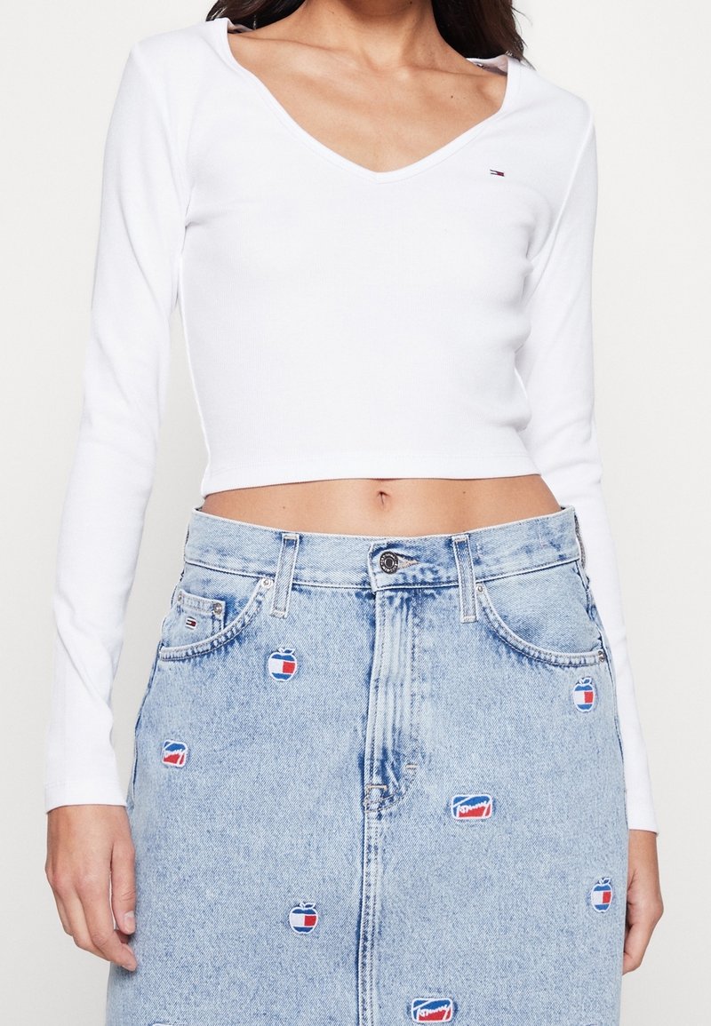 Weißes langärmliges Oberteil mit V-Ausschnitt, kombiniert mit einem hellblauen Jeansrock, der bunte, bestickte Logo-Patches gleichmäßig verteilt trägt.