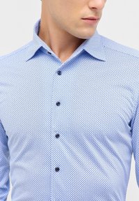 Camicia azzurro chiaro con un piccolo motivo geometrico, colletto strutturato e bottoni neri, che mette in mostra una trama di tessuto liscia.