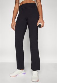 Personne portant un pantalon noir taille haute et des baskets blanches avec des accents violets, tenant un objet rond blanc dans la main droite.
