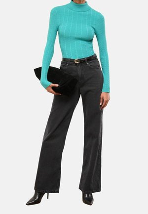 Personne portant un col roulé en maille torsadée turquoise, un jean noir à jambes larges avec ceinture, des talons noirs pointus, tenant une pochette noire.