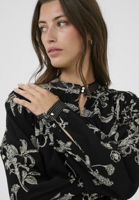 Schwarze Bluse mit filigraner weißer Blumenstickerei, hohem Kragen mit Knopfdetails und rüschigen Ballonärmeln mit kontrastierenden Manschetten.