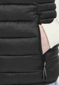 Gilet noir matelassé avec des coutures verticales et une poche latérale, présentant une fermeture éclair avec des tirettes doubles et une manche côtelée blanche en dessous.