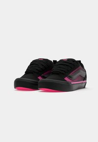 Et par svarte og rosa lave sneakers med svarte lisser og et bølgedesign på sidene, plassert mot en hvit bakgrunn.