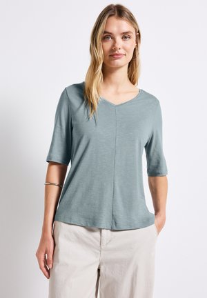 Camicia azzurra chiara a V con maniche corte, realizzata in tessuto morbido. Presenta un dettaglio di cucitura frontale e una vestibilità rilassata. Abbinata a pantaloni chiari.