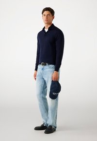 Marineblaues Langarm-Polohemd, hellblaue Jeans, schwarze Slipper. Hält eine Marineblau-Basecap mit weißem Text. Einfaches, lässiges Outfit.