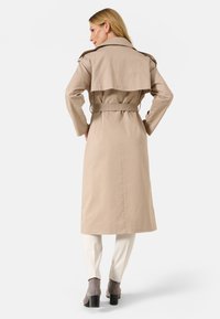 Cappotto trench beige con una parte posteriore fluida, vita cintata e alette sulle spalle. Abbinato a pantaloni crema a fantasia e stivali grigi con tacco.