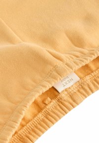 Gula bomullsbebisbyxor med elastiskt midjeband och mjuk textur. Etikett med varumärkesnamn synlig. Enkel design, utan mönster.
