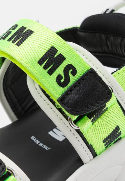 Sandal com tiras entrelaçadas em preto e verde neon, apresentando texto impresso e detalhes em couro, além de uma sola branca. "FABRICADO NA ITÁLIA" visível no interior.