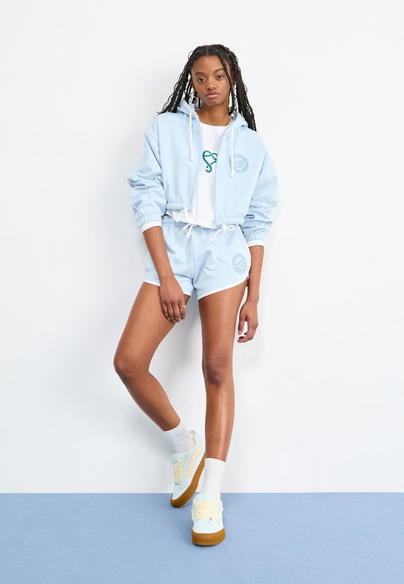 Haut cropped bleu clair avec cordons, shorts assortis et baskets pastel. Les designs comprennent des logos à la fois sur le haut et sur les shorts.