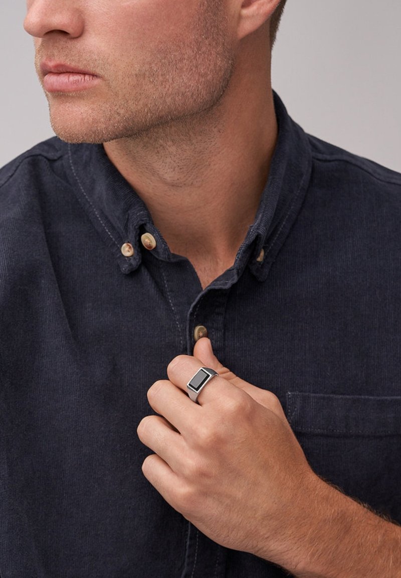 Bague rectangulaire noire avec un band en argent portée à la main droite. Le modèle porte une chemise boutonnée sombre avec une finition en tissu texturé.