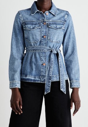 Person iført en blå denimjakke med knapper og brystlommer samt et bindebælte i taljen, kombineret med sorte bukser.