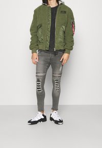 Chaqueta bomber verde oliva con textura de forro polar y detalles en satén, combinada con jeans grises desgastados y zapatillas deportivas en blanco y negro.
