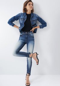 Chaqueta de mezclilla con dos bolsillos en el pecho, combinada con unos vaqueros de tiro alto y azul desgastado, y tacones negros con tiras. Textura suave y corte clásico.