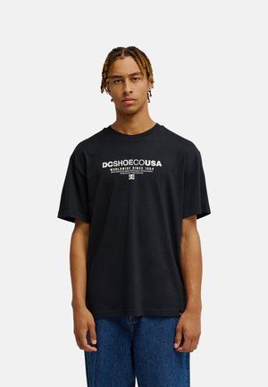 Młody mężczyzna z krótkimi warkoczykami ma na sobie luźny czarny t-shirt DC SHOES USA z białym napisem i niebieskie dżinsy, stojąc na tle jednolitego jasnego tła.