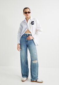 Camisa a rayas blanca y azul con logo, vaqueros anchos de mezclilla desgastados de tiro medio en azul claro, y zapatillas blancas con detalles en negro. Gafas de sol negras.
