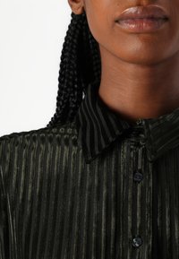 Camisa verde escura com riscas verticais pretas, gola com colarinho e placket de botões visível; feita de tecido texturizado.