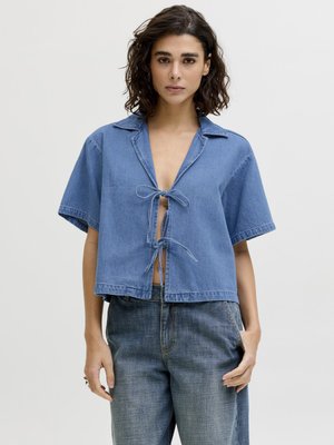 Mladá osoba s kučeravými vlasmi nosí modrú rifľovú košeľu s krátkym rukávom, zviazanú vpredu, a voľné rifle s vysokým pásom, stojaca proti bielemu pozadiu.