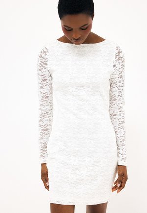 Kobieta w dopasowanej białej koronkowej sukience z długimi przeźroczystymi koronkowymi rękawami, patrząca w dół na jednolitym białym tle.