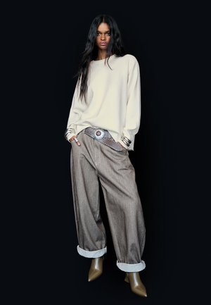 Licht beige oversized trui, grijze wijde broek met omgeslagen zoom, zilveren gespschoen, en beige puntige hakken.