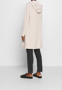 Cardigan long beige clair avec capuche, texture côtelée, fentes sur les côtés, porté sur un jean sombre et des sandales noires, vu de dos.