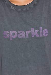 Grå T-shirt med rund halsudskæring og et skinnende lilla "sparkle" tekstdesign på forsiden. Blødt stof med en struktureret overflade.