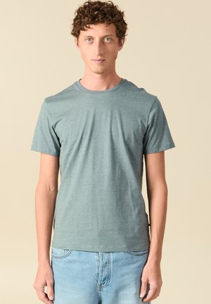 Giovane uomo con capelli ricci che indossa una maglietta grigia a maniche corte semplice e jeans azzurri, in piedi davanti a uno sfondo beige.