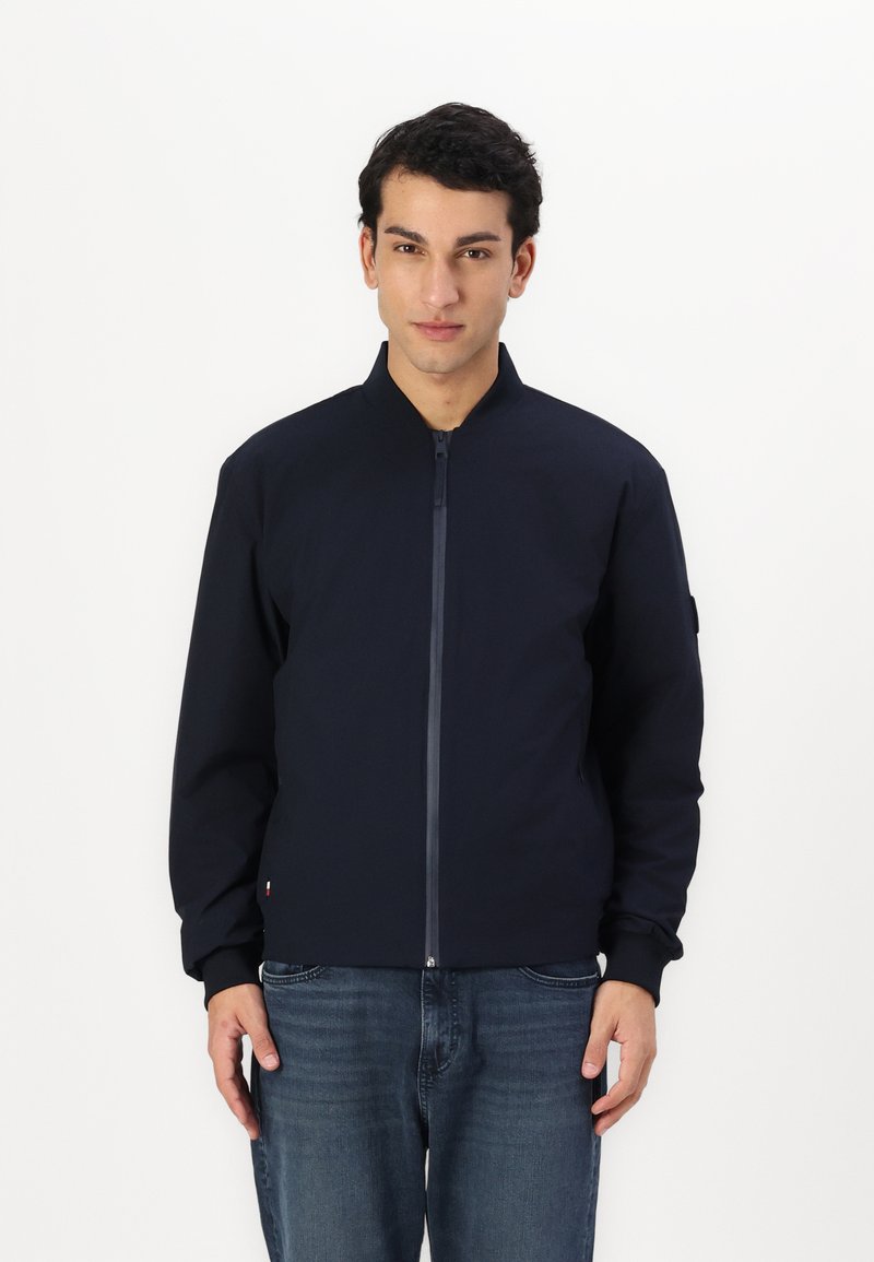 Giacca bomber navy con chiusura a zip, polsini e colletto a coste. Realizzata in tessuto liscio, presenta una patch con logo sulla manica.