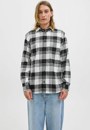 Ung person med skulderlangt hår iført en sort og hvid ternet skjorte med knapper og lyseblå vide jeans, står mod en hvid baggrund.