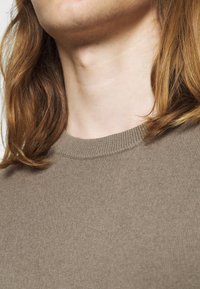 Gros plan sur le cou et le bas du visage d'une personne à la peau claire et aux cheveux châtain roux portant un pull en maille marron.
