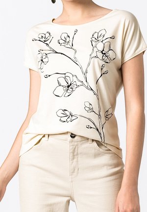 T-shirt beige clair avec un design de contour floral noir, manches courtes, décolleté arrondi, coupe décontractée. Assorti avec un pantalon de couleur claire.