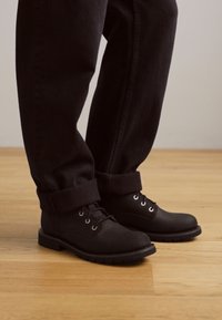 Botas de cuero negras con punta redonda, diseño con cordones y ojales de metal, combinadas con pantalones de mezclilla negros enrollados sobre un suelo de madera.