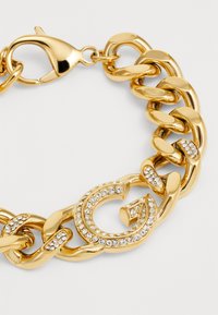 Gouden kettingarmband met grote schakels, een karabijnsluiting en een centrale "G" versierd met heldere rhinestones. Zachte textuur, glanzende afwerking.