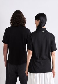 Dos personas están de pie de espaldas, vistiendo camisetas negras de manga corta; una lleva jeans negros y la otra una falda blanca con pliegues.