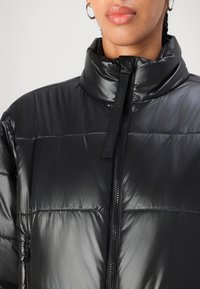 Czarna kurtka puchowa z wysokim kołnierzem i zamkiem błyskawicznym z przodu. Wykonana z błyszczącego materiału z subtelnym poziomym przeszyciem. Widoczne logo marki.