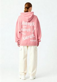 Felpa con cappuccio rosa con testo bianco "AMOUR", taglio oversize, indossata con pantaloni cargo color crema e sneakers bianche massicce.