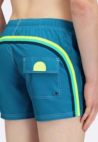 Pantaloni da bagno turchesi con strisce giallo neon e navy. Presentano una tasca laterale con un patch circolare del logo. Tessuto leggero e ad asciugatura rapida.