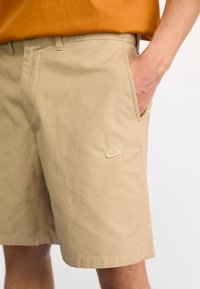 Mies, joka käyttää beigejä Nike-shortsseja ja jolla on käsi taskussa, yhdistettynä oranssiin paitaaan valkoista taustaa vasten.