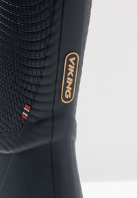 Gros plan sur une botte viking noire montrant un panneau latéral texturé et un logo ovale avec bordure et lettrage dorés.