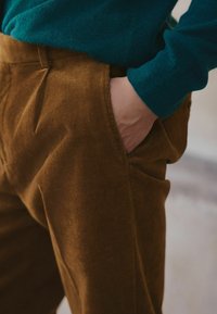 Pantaloni din țesătură de catifea maro, cu buzunare laterale, asortați cu un pulover teal. Textura de catifea este vizibilă, evidențiind designul său cu pliuri.