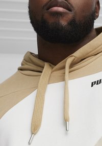 Beżowa i biała bluza z kapturem z sznurkiem, wykonana z teksturowanego materiału, z czarnym logo "PUMA" na klatce piersiowej.