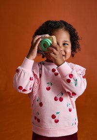 Rosa Sweatshirt mit Kirschdruck, Spitzenelementen an den Ärmeln, kombiniert mit einer Holzspielzeugkamera, vor einem orangen Hintergrund.