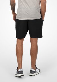 Shorts de sport noirs en tissu texturé, avec des poches latérales, associés à des chaussures de sport grises et blanches. Arrière-plan neutre.